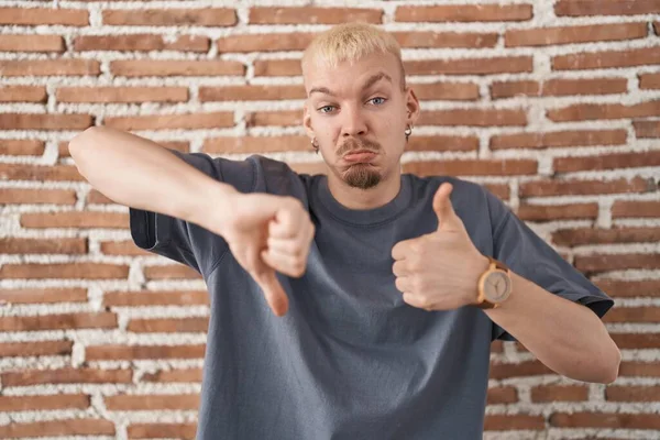 Young caucasian man standing over bricks wall doing thumbs up and down, disagreement and agreement expression. crazy conflict