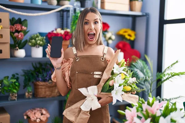 Young woman working at florist shop showing smartphone screen celebrating crazy and amazed for success with open eyes screaming excited.