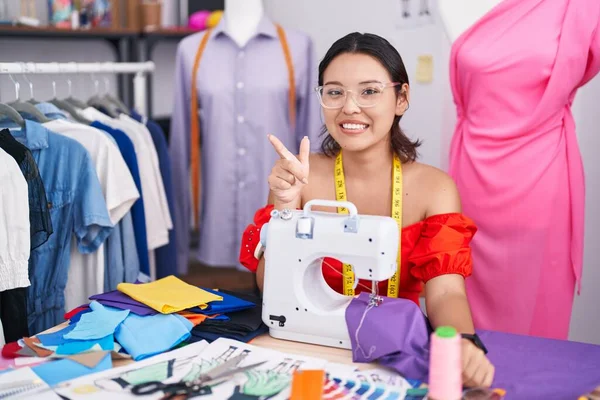 Hispanic young woman dressmaker designer using sewing machine cheerful with a smile on face pointing with hand and finger up to the side with happy and natural expression