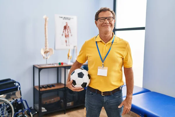 Senior man working at sport physiotherapy clinic looking positive and happy standing and smiling with a confident smile showing teeth