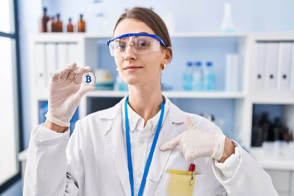 Young caucasian woman working at scientist laboratory holding bitcoin pointing finger to one self smiling happy and proud