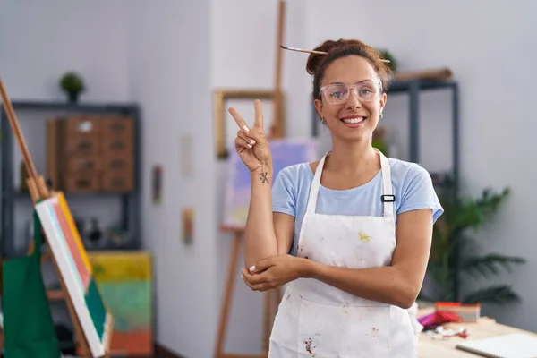 Brunette woman painting at art studio smiling with happy face winking at the camera doing victory sign. number two.