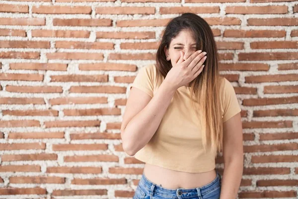 若いブルネットの女性がレンガの壁の上に立って何か臭いと嫌な 耐えられない匂いを嗅ぎ 鼻の上に指で息を保持する 臭いが悪い — ストック写真