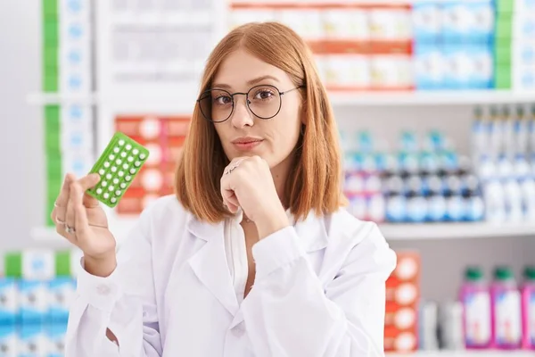 若い赤毛の女性は薬局で働いています避妊薬を保持しています深刻な顔あごに手で質問について考えて 混乱のアイデアについて思慮深く — ストック写真