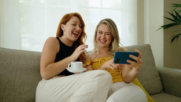 Two women drinking coffee watching video on smartphone at home