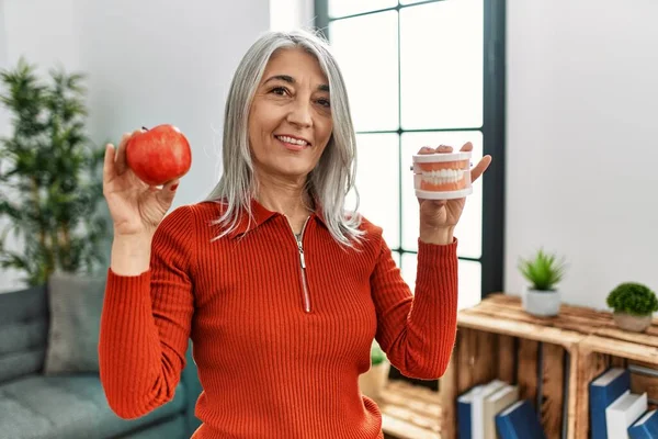 Middle age grey-haired woman smiling confident holding red apple and denture at home
