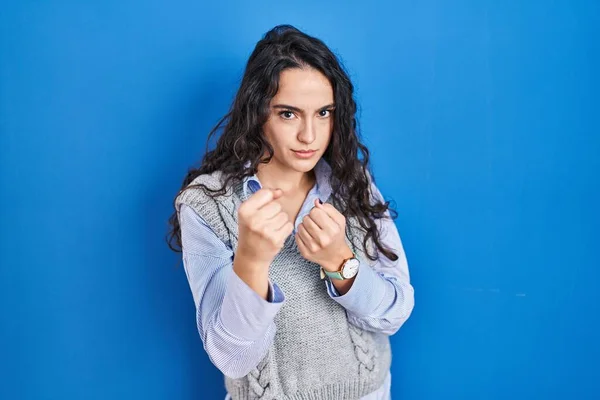 Young brunette woman standing over blue background ready to fight with fist defense gesture, angry and upset face, afraid of problem