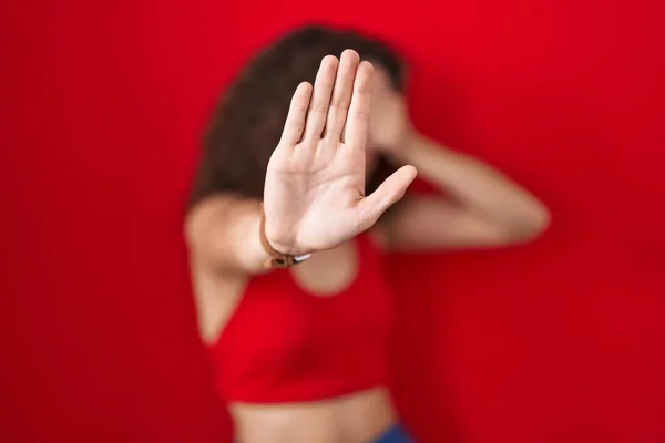 Hispanic woman with curly hair standing over red background covering eyes with hands and doing stop gesture with sad and fear expression. embarrassed and negative concept.
