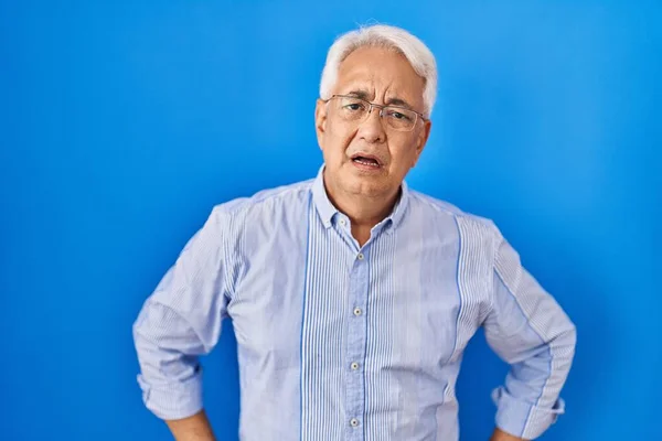 Hispanic senior man wearing glasses in shock face, looking skeptical and sarcastic, surprised with open mouth