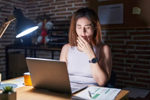 Brunette woman working at the office at night bored yawning tired covering mouth with hand. restless and sleepiness.