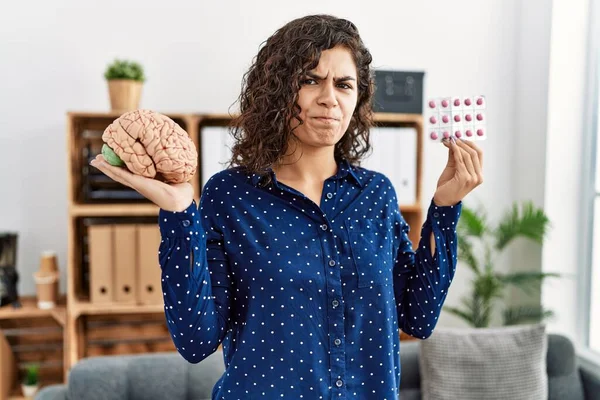 Young brunette woman holding brain and pills as mental health concept skeptic and nervous, frowning upset because of problem. negative person.