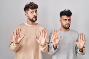 Young homosexual couple standing over white background moving away hands palms showing refusal and denial with afraid and disgusting expression. stop and forbidden. 