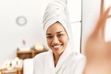 Bornoz giyen genç Latin kadın güzellik merkezindeki kameranın önünde selfie çekiyor.