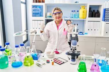 Young hispanic woman working at scientist laboratory doing make up pointing thumb up to the side smiling happy with open mouth 