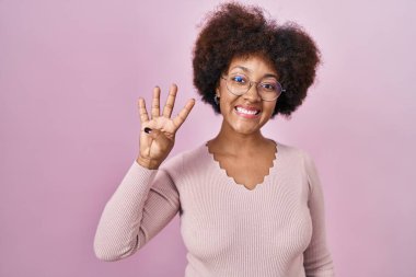 Genç Afrikalı Amerikalı kadın pembe arka planda duruyor ve parmaklarını dört numaraya doğrultuyor ve gülümsüyor kendinden emin ve mutlu.. 