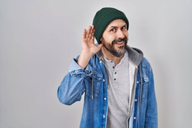 Young hispanic man with tattoos wearing wool cap smiling with hand over ear listening an hearing to rumor or gossip. deafness concept. 