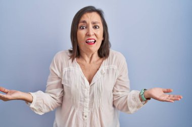 Orta yaşlı Latin bir kadın mavi arka planda duruyor. Kolları açık ve kafası karışık, hiçbir fikri yok.. 