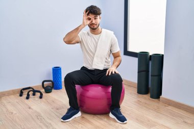 Hispanic man with beard sitting on pilate balls at yoga room doing ok gesture with hand smiling, eye looking through fingers with happy face. 