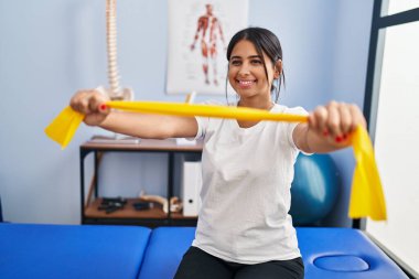 Genç Latin kadın hasta fizik tedavi kliniğinde elastik bant kullanarak rehabilitasyon seansı yapıyor.