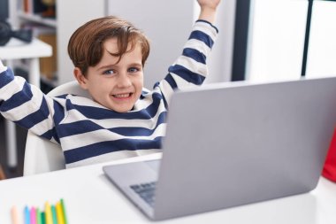 Adorable hispanic boy student using laptop with cheerful expression at classroom