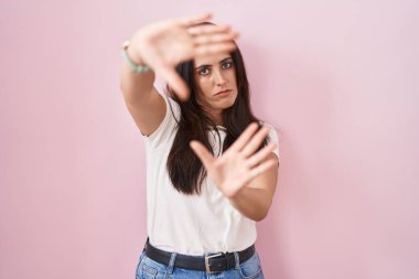 Young brunette woman standing over pink background doing frame using hands palms and fingers, camera perspective 