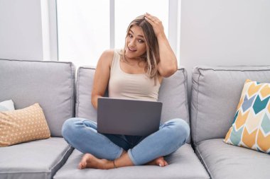 Blonde caucasian woman using laptop at home sitting on the sofa stressed and frustrated with hand on head, surprised and angry face 