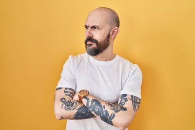 Young hispanic man with tattoos standing over yellow background looking to the side with arms crossed convinced and confident 