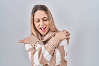 Young blonde woman wearing turtleneck sweater over isolated background hugging oneself happy and positive, smiling confident. self love and self care 