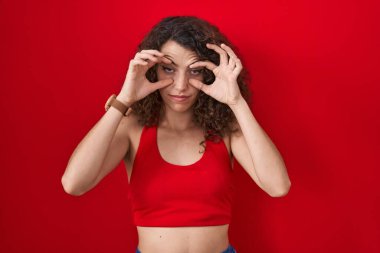 Hispanic woman with curly hair standing over red background trying to open eyes with fingers, sleepy and tired for morning fatigue 