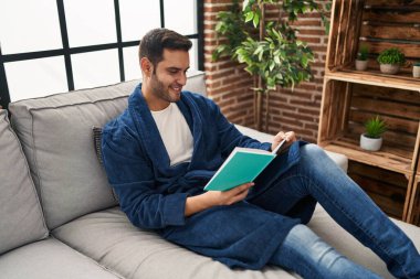 Evde bornoz giyen genç İspanyol adam kitap okuyor.