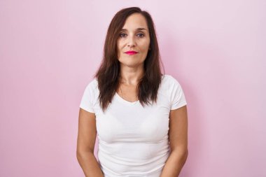 Middle age brunette woman standing over pink background relaxed with serious expression on face. simple and natural looking at the camera. 