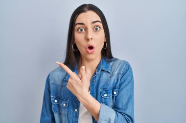 Hispanic woman standing over blue background surprised pointing with finger to the side, open mouth amazed expression. 