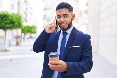 Young latin man business worker talking on smartphone drinking coffee at street