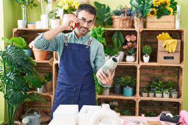 Young hispanic man florist talking on smartphone holding plant pot at flower shop