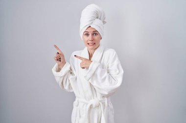 Blonde caucasian woman wearing bathrobe pointing aside worried and nervous with both hands, concerned and surprised expression 