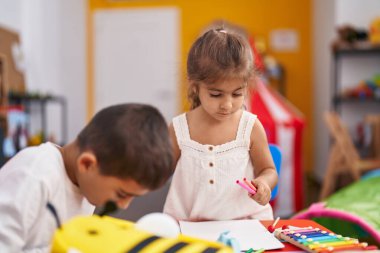 Erkek ve kız kardeş öğrencileri anaokulunda masaya oturup kağıt çiziyorlar.