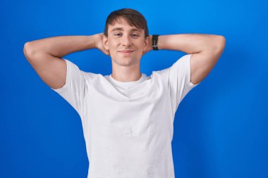 Caucasian blond man standing over blue background relaxing and stretching, arms and hands behind head and neck smiling happy 