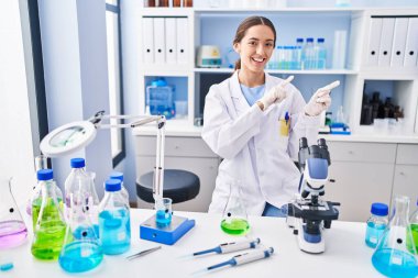 Young brunette woman working at scientist laboratory smiling and looking at the camera pointing with two hands and fingers to the side. 