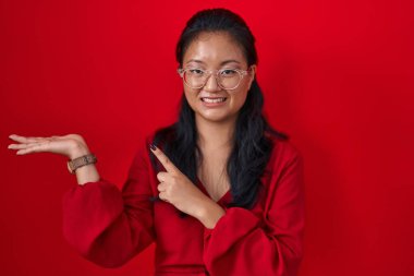 Asian young woman standing over red background amazed and smiling to the camera while presenting with hand and pointing with finger. 