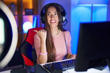 Young hispanic woman playing video games smiling with happy face looking and pointing to the side with thumb up. 