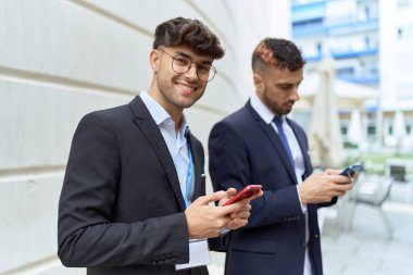 İki İspanyol işadamı sokakta akıllı telefon kullanarak gülümsüyor.