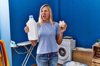 Beautiful woman doing laundry holding detergent bottle and piggy bank angry and mad screaming frustrated and furious, shouting with anger. rage and aggressive concept. 