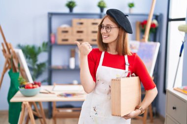 Young redhead woman at art studio holding art case smiling with happy face looking and pointing to the side with thumb up. 