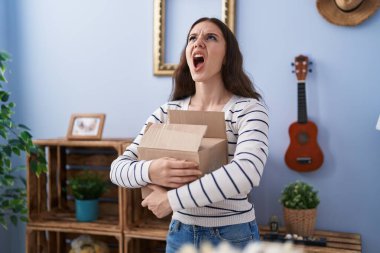 Young hispanic girl opening cardboard box angry and mad screaming frustrated and furious, shouting with anger looking up. 