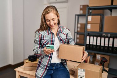 Genç sarışın kadın ecommerce iş işçisi ofiste akıllı telefon paketi kullanıyor.