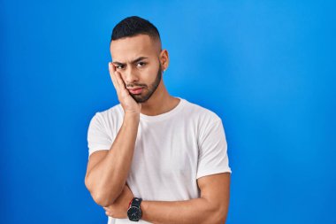 Young hispanic man standing over blue background thinking looking tired and bored with depression problems with crossed arms. 