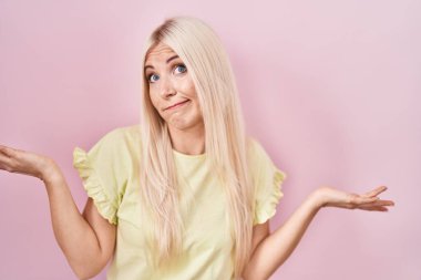 Caucasian woman standing over pink background clueless and confused expression with arms and hands raised. doubt concept. 