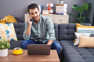 Young hispanic man talking on smartphone using laptop at home