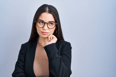 Young brunette woman standing over blue background with hand on chin thinking about question, pensive expression. smiling with thoughtful face. doubt concept. 