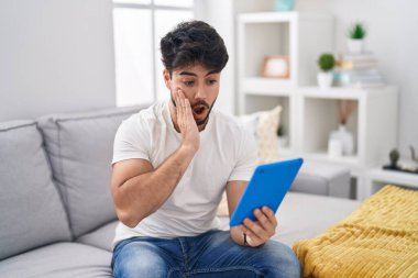 Hispanic man with beard using touchpad sitting on the sofa afraid and shocked, surprise and amazed expression with hands on face 